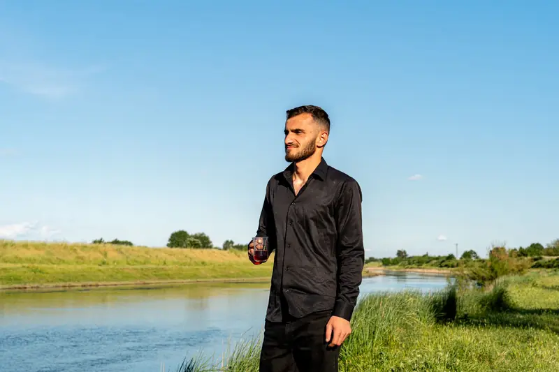 Man in a black shirt stands by a river enjoying nature under a clear blue sky