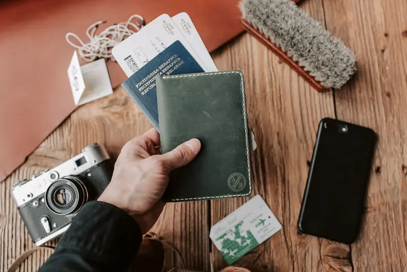 Close-up of a hand holding a passport with travel essentials including a camera, phone, and tickets on a wooden table